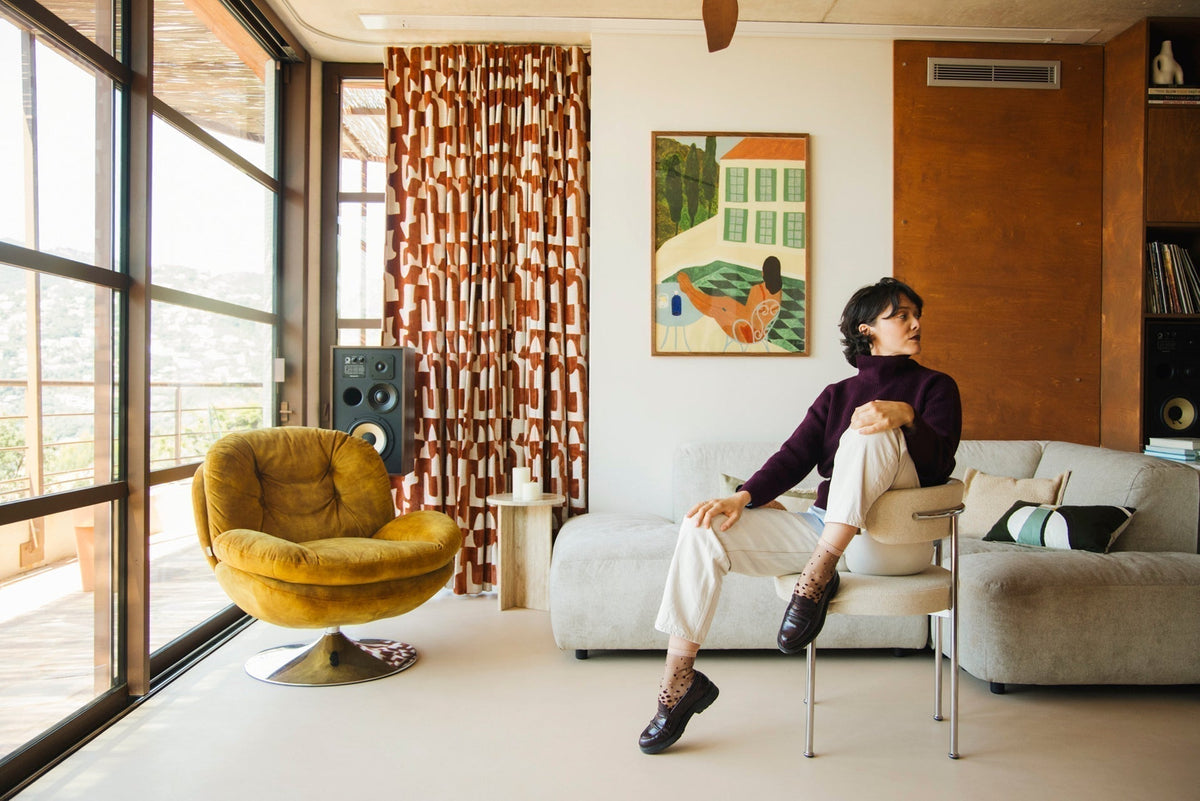 Mannequin qui porte des chaussettes transparentes au dégradé de poids bordeaux et doré, vêtue d'un pantalon beige, pull bordeaux et de mocassins noirs. Elle est assise sur une chaise beige, dans un salon au style rétro année 70, et regarde vers la droite.