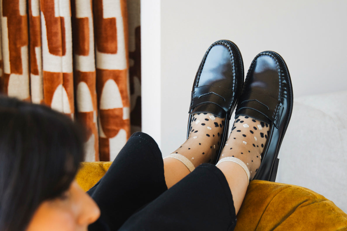 Zoom sur les pieds d'une mannequin portant des chaussettes transparentes au dégradé de pois noir et doré, vêtue d'un pantalon noir et de mocassins noirs. La femme est assise sur un fauteuil jaune moutarde, les pieds en l'air.