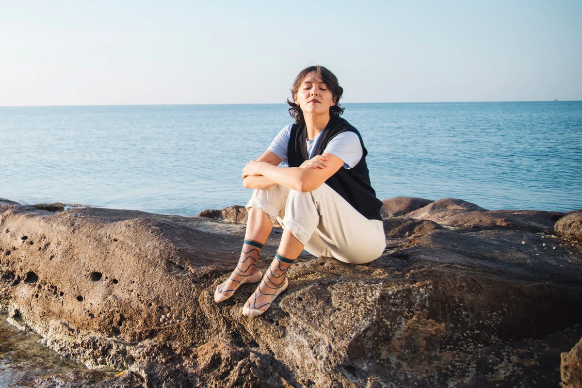 Femme portant des chaussettes transparentes aux courbes bleues, vêtue d'un pantalon crème, d'un tee-shirt bleu et d'une veste sans manche bleue foncé, assise sur des rochers au bord de la mer.