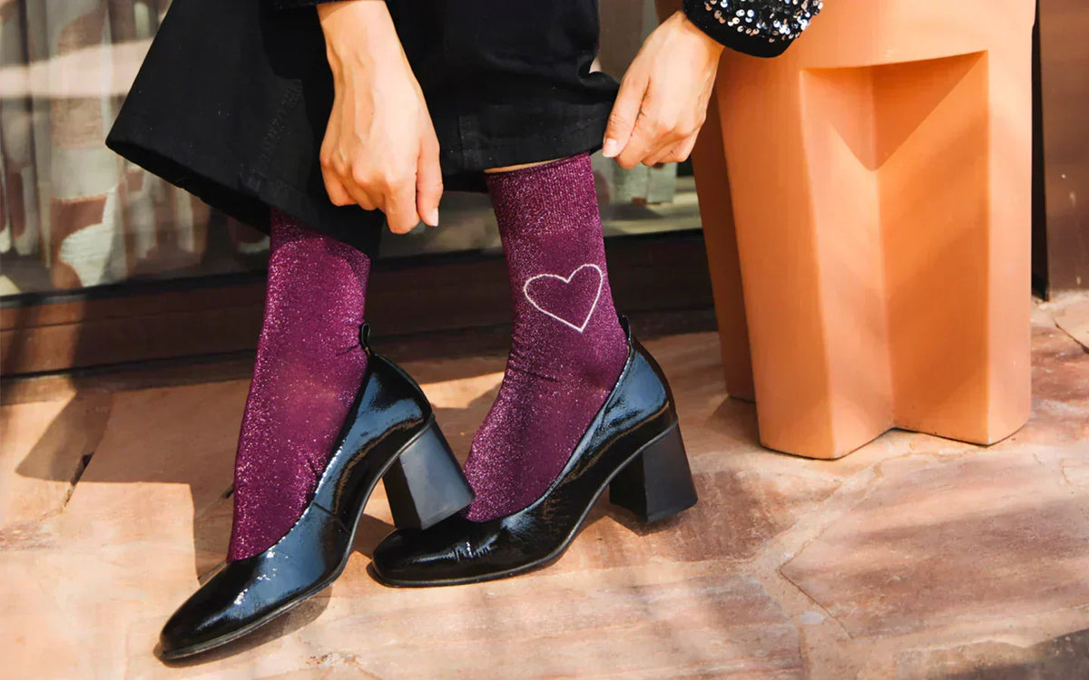 Zoom sur les pieds d’une mannequin portant des chaussettes en coton pailleté violet et bordeaux, décorées d’un cœur au contour blanc sur la cheville externe gauche.  