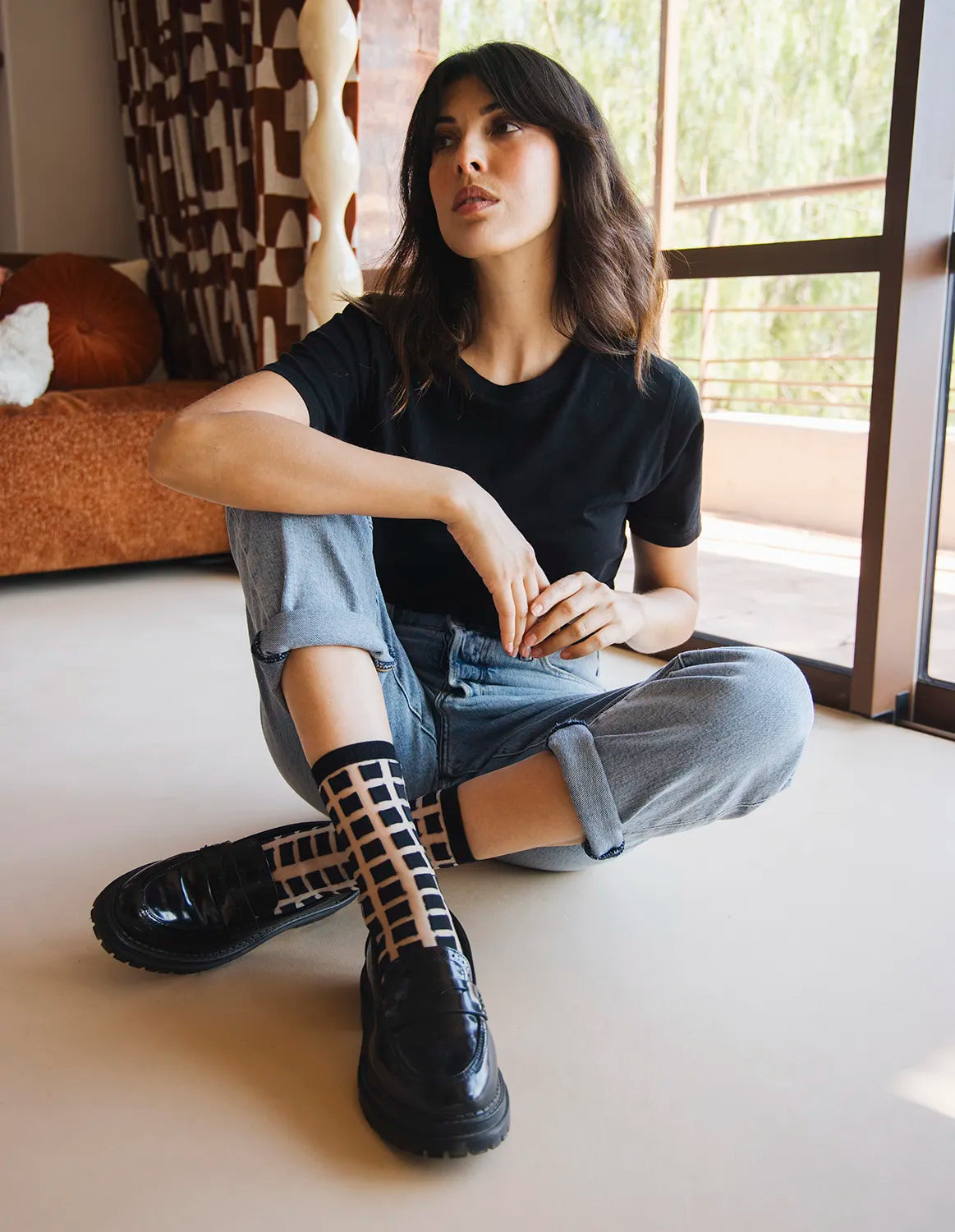 femme assise par terre avec jean bleu tshirt noir et chaussettes transparentes à carreaux noirs jambes croisées ambiance décontractée