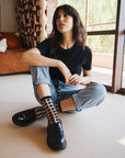femme assise par terre avec jean bleu tshirt noir et chaussettes transparentes à carreaux noirs jambes croisées ambiance décontractée