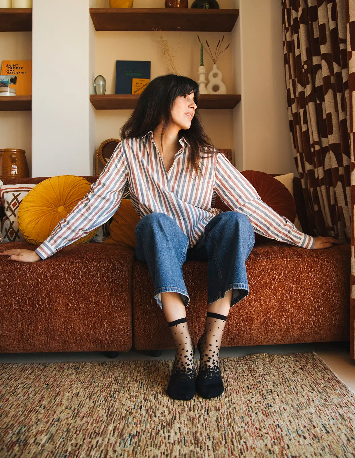 Chaussettes transparentes à pois noirs, portées par une femme assise sur un canapé portant un jean bleu et une chemise rayée dans une architecture moderne et 70's